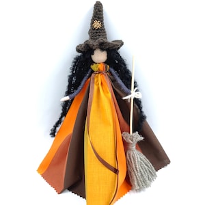 May include: A handmade witch doll with long black yarn hair, wearing a brown crocheted hat with a spider embellishment, a multi-colored fabric dress, and holding a wooden stick with a gray yarn broom.