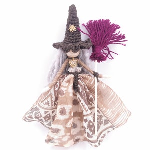 May include: A handmade witch doll with a brown crocheted hat, a long white yarn wig, a brown and white patterned fabric dress, and a purple yarn broom.