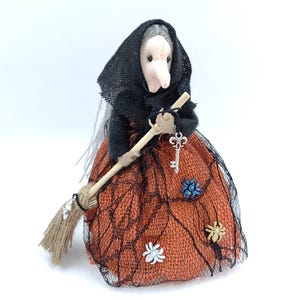 Può includere: Bambola di strega fatta a mano con un velo nero e un vestito arancione. La bambola tiene in mano una scopa e un portachiavi a forma di chiave. L'abito è decorato con una rete nera e piccoli abbellimenti floreali. Decorazione di Halloween.