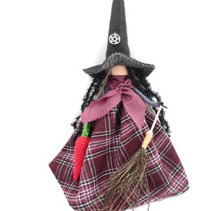 Può includere: Una bambola di strega fatta a mano con un cappello nero a punta, un vestito a quadri bordeaux, una carota rossa all'uncinetto e una scopa marrone.