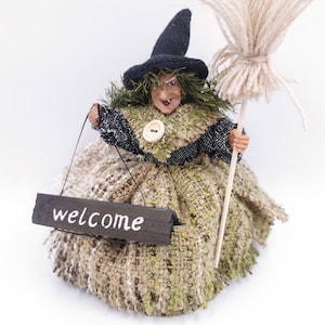 May include: A witch doll wearing a black hat and a green and brown dress, holding a broom and a sign that says "welcome".