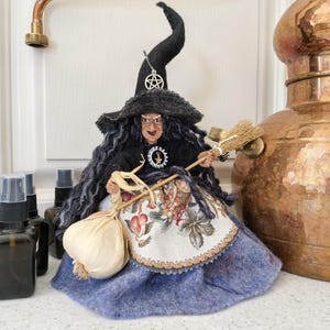 Puede incluir: Una muñeca bruja artesanal con cabello largo y oscuro, sombrero negro puntiagudo con un dije de pentáculo plateado y blusa negra. La muñeca viste un delantal con estampado floral sobre una falda azul y sostiene una escoba y ajo seco.