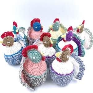 May include: A collection of handmade crochet rooster figurines. Each rooster features a unique color scheme, including red, blue, pink, and purple, with detailed combs, beaks, and tail feathers. The figurines are arranged on a white background.