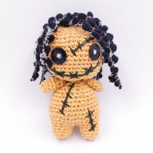 Puede incluir: Una muñeca vudú de crochet con pelo de hilo negro, ojos cosidos y boca cosida. La muñeca lleva una sonrisa cosida y tiene una cicatriz cosida en el pecho.