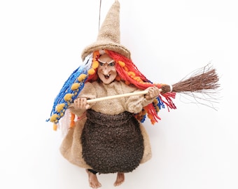 May include: A whimsical witch doll with a burlap hat and dress, holding a broom. The doll has colorful yarn hair in shades of red, blue, and orange. The witch's face has a mischievous expression, perfect for Halloween decor.
