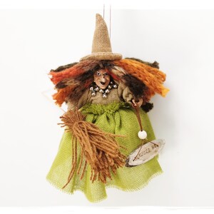 May include: A handmade witch doll with a brown burlap hat, orange and brown yarn hair, a green burlap skirt, and a brown yarn broom. The doll is holding a wooden sign that says "Blessed Be".