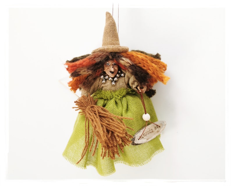May include: A handmade witch doll with a brown burlap hat, orange and brown yarn hair, a green burlap skirt, and a brown yarn broom. The doll is holding a wooden sign that says "Blessed Be".