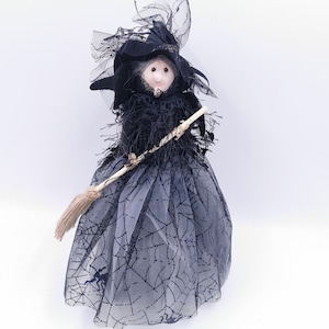 Può includere: Una bambola di strega nera con un cappello nero e una scopa. La bambola indossa un vestito nero con un motivo a ragnatela.