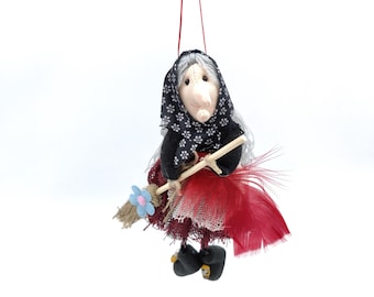 Può includere: Un ornamento di bambola di strega fatto a mano con un vestito a motivi bianco e nero, una gonna rossa e una scopa con un fiore blu. La bambola ha una parrucca bianca e un naso lungo.