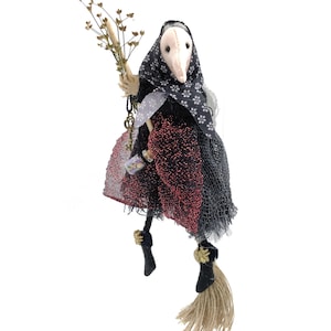 Könnte beinhalten: Handgemachte Hexenpuppe mit weißer schnabelartiger Maske, schwarzer Blumenkopfbedeckung und rotem und schwarzem Kleid. Die Puppe hält ein Bündel getrockneter Blumen und eine kleine Flasche. Sie schwebt in der Luft.