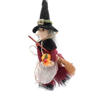 Puede incluir: Muñeca de bruja de Halloween con sombrero negro, vestido rojo y negro y escoba. La bruja tiene pelo gris y sostiene una calabaza con una mariquita. La muñeca es de tela y tiene un aspecto rústico.