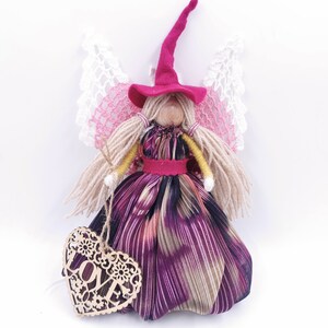 May include: A handmade fairy doll with pink and white wings, a red felt hat, and a long purple and brown dress. The doll is holding a wooden heart with the word "LOVE" carved into it.