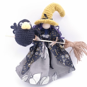 May include: A crocheted witch doll with a yellow hat and a black crow perched on her broom. She is wearing a black and grey dress with a floral pattern.