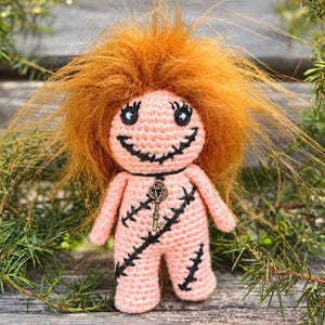 Può includere: Una bambola all'uncinetto con pelle in filato rosa chiaro, capelli arancioni vivaci e spettinati e occhi a bottone neri. La bambola presenta una bocca nera cucita e motivi a punto croce neri sul corpo, con una piccola chiave in bronzo al collo.