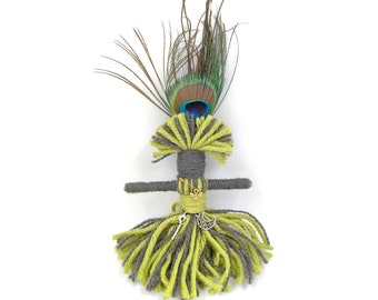Op de afbeelding: Een grijze en groene garenpop met een pauwenveer op zijn hoofd. De pop heeft een sleutel en een halve maan hanger aan zijn taille.