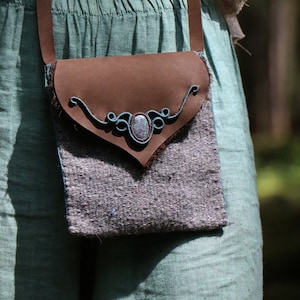 May include: A brown leather crossbody bag with a grey fabric flap and a turquoise gemstone accent. The bag is worn over a light blue linen top and matching skirt.