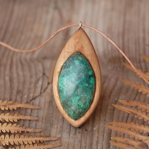 May include: A marquise-shaped pendant featuring a smooth, mottled green stone set within a light brown wooden frame. The pendant is suspended from a thin, light-colored cord, creating a natural aesthetic with its organic shape and earthy tones.