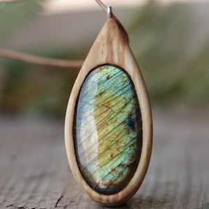 May include: A teardrop-shaped pendant featuring a polished labradorite gemstone with iridescent blue and yellow flashes, encased in a smooth, light-colored wooden frame. The pendant hangs from a thin cord attached to a small metal bail.