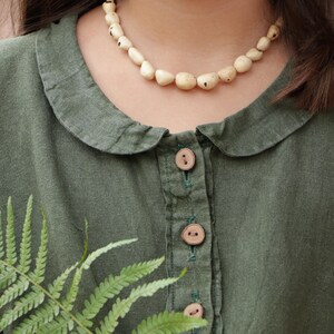 May include: An olive green long-sleeved shirt featuring a rounded Peter Pan collar, a front button placket with three light brown wooden buttons, and a necklace made of irregular, cream-colored, speckled beads worn around the neck.