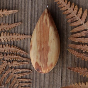 May include: A smooth, teardrop-shaped wooden pendant featuring a natural grain pattern with light tan and reddish-brown streaks. The pendant has a small silver-toned metal bail attached to the top point, allowing it to be suspended as a necklace.
