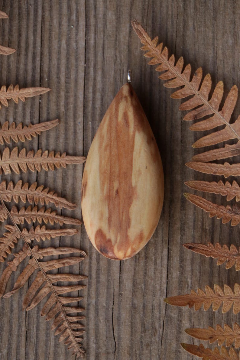 May include: A smooth, teardrop-shaped wooden pendant featuring a natural grain pattern with light tan and reddish-brown streaks. The pendant has a small silver-toned metal bail attached to the top point, allowing it to be suspended as a necklace.