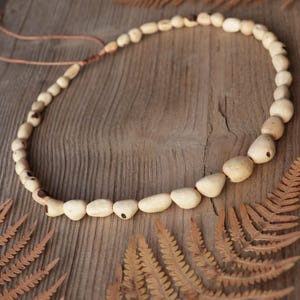 May include: A necklace featuring a series of smooth, light beige, irregularly shaped wooden beads strung on a thin cord. The beads vary in size, with larger, more prominent pieces positioned along the lower curve, and the cord includes a knot.