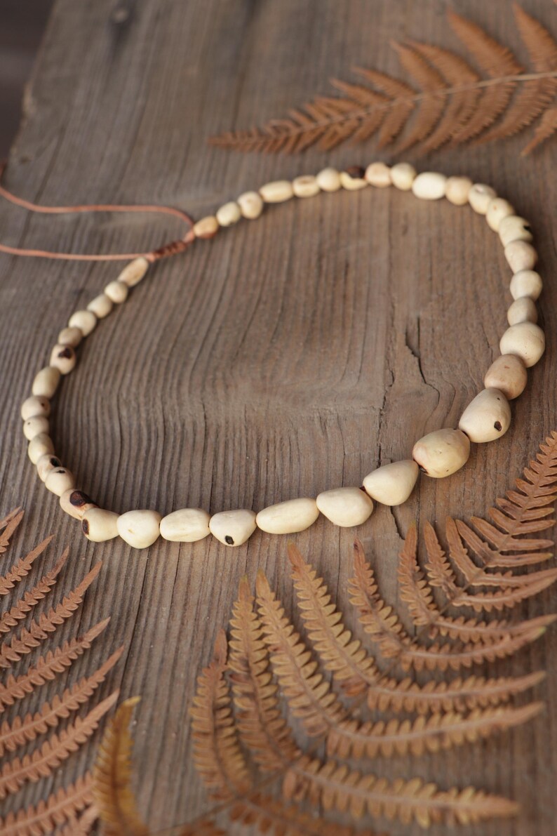 May include: A necklace featuring a series of smooth, light beige, irregularly shaped wooden beads strung on a thin cord. The beads vary in size, with larger, more prominent pieces positioned along the lower curve, and the cord includes a knot.