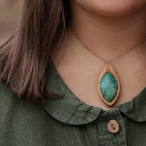 May include: A necklace featuring a marquise-shaped pendant with a light brown wooden frame surrounding a textured, mottled green stone inlay. The pendant hangs from a thin, light brown cord that rests against the neck.