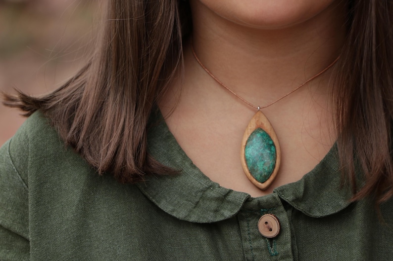 May include: A necklace featuring a marquise-shaped pendant with a light brown wooden frame surrounding a textured, mottled green stone inlay. The pendant hangs from a thin, light brown cord that rests against the neck.