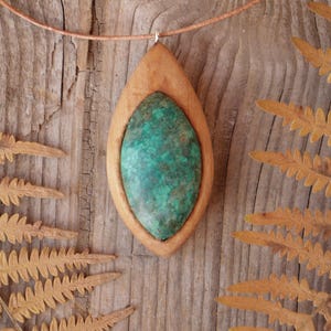 May include: A pendant necklace featuring a marquise-shaped green and brown mottled stone set within a smooth, light-toned wooden frame. The pendant is suspended from a thin, light brown cord that circles the top of the frame.