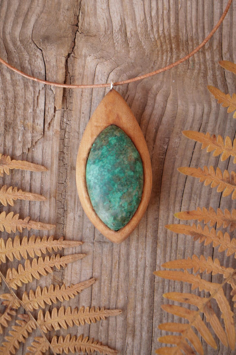 May include: A pendant necklace featuring a marquise-shaped green and brown mottled stone set within a smooth, light-toned wooden frame. The pendant is suspended from a thin, light brown cord that circles the top of the frame.