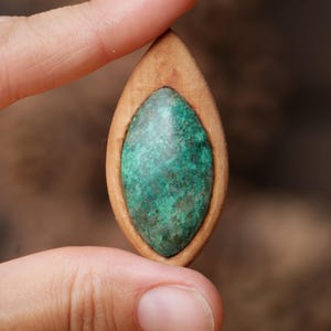 May include: A marquise-shaped pendant featuring a smooth, light brown wooden frame surrounding a textured, mottled green stone inlay. The stone displays variations of dark and light green hues with a natural, uneven surface finish.