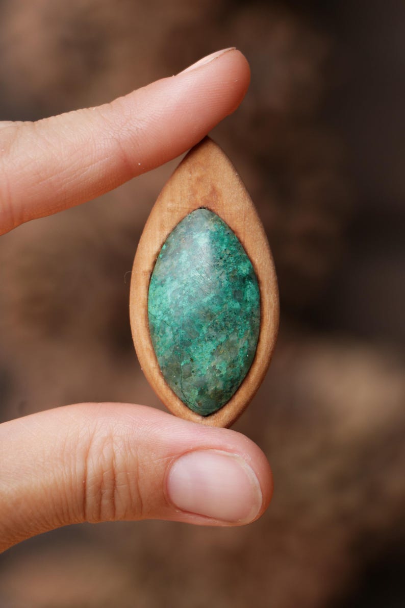 May include: A marquise-shaped pendant featuring a smooth, light brown wooden frame surrounding a textured, mottled green stone inlay. The stone displays variations of dark and light green hues with a natural, uneven surface finish.