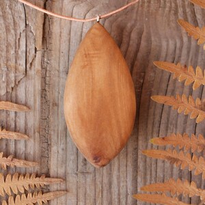 May include: A smooth, light brown wooden pendant shaped like an elongated almond or leaf. The pendant is suspended from a thin, light-colored cord. The wood grain is visible, showing natural variations in color and texture across the surface.