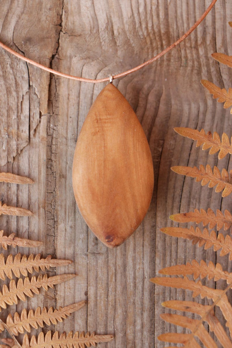 May include: A smooth, light brown wooden pendant shaped like an elongated almond or leaf. The pendant is suspended from a thin, light-colored cord. The wood grain is visible, showing natural variations in color and texture across the surface.