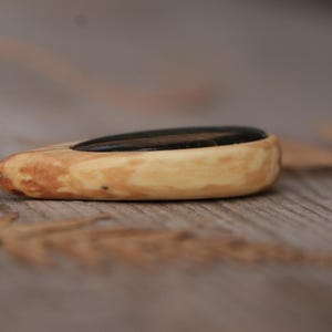 May include: A teardrop-shaped pendant featuring a smooth, light-colored wood frame with a dark, polished stone inlay. The pendant is attached to a thin, light-colored cord by a small silver-toned metal jump ring, creating a simple necklace design.