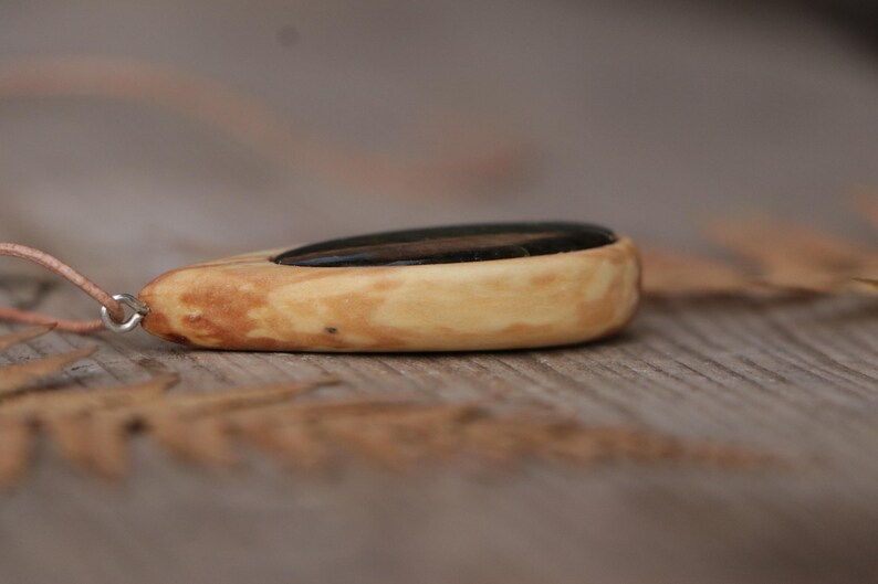 May include: A teardrop-shaped pendant featuring a smooth, light-colored wood frame with a dark, polished stone inlay. The pendant is attached to a thin, light-colored cord by a small silver-toned metal jump ring, creating a simple necklace design.