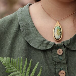 May include: A teardrop-shaped pendant featuring a polished, iridescent oval stone set within a light-colored wooden frame. The pendant hangs from a thin, brown cord. The piece is worn over a dark olive green collared shirt with wooden buttons.