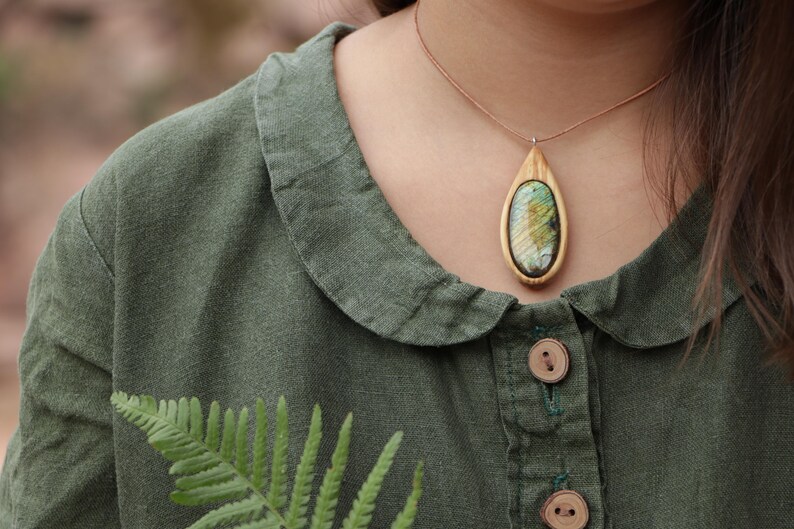 May include: A teardrop-shaped pendant featuring a polished, iridescent oval stone set within a light-colored wooden frame. The pendant hangs from a thin, brown cord. The piece is worn over a dark olive green collared shirt with wooden buttons.