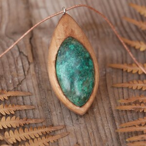 May include: A pendant featuring a smooth, marquise-shaped green stone with dark inclusions, set within a light-colored wooden frame. The pendant is suspended from a thin, light brown cord, creating a simple and natural aesthetic for this jewelry piece.