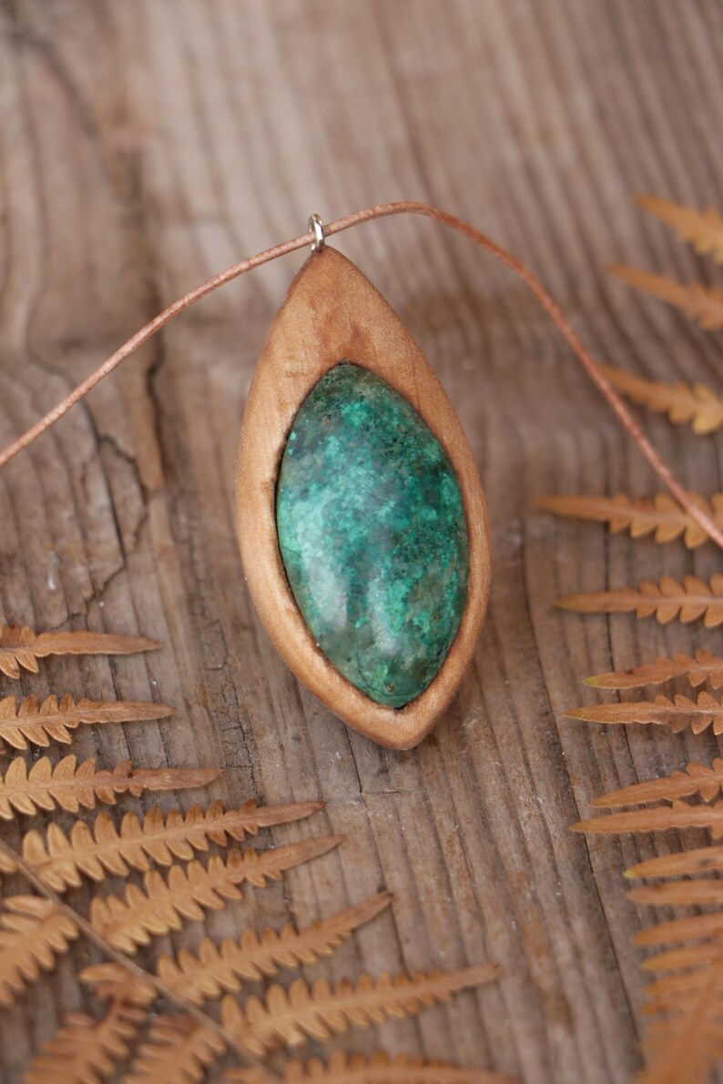 May include: A pendant featuring a smooth, marquise-shaped green stone with dark inclusions, set within a light-colored wooden frame. The pendant is suspended from a thin, light brown cord, creating a simple and natural aesthetic for this jewelry piece.