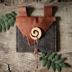 May include: A small brown leather pouch with a gray felt lining and a wooden spiral charm. The pouch has a leather strap and is sitting on a wooden surface.