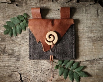 May include: A small brown leather pouch with a gray felt lining and a wooden spiral charm. The pouch has a leather strap and is sitting on a wooden surface.