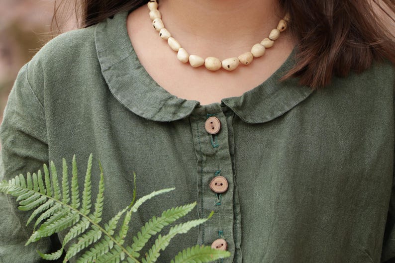 May include: An olive green long-sleeved shirt featuring a rounded Peter Pan collar, a front button placket with three light brown wooden buttons, and a necklace made of irregular, cream-colored, speckled beads worn around the neck.