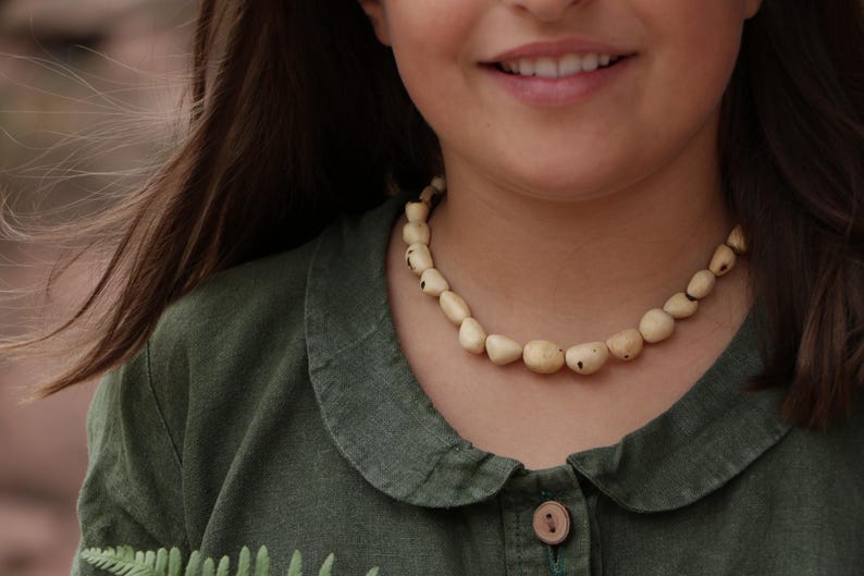May include: A necklace composed of smooth, irregularly shaped, off-white beads with small dark brown inclusions. The beads are strung together in a single strand, resting around the neck, featuring a natural, matte finish and varied organic forms.