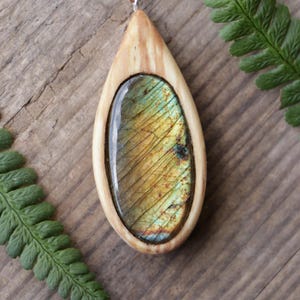 May include: A teardrop-shaped pendant featuring a polished, iridescent labradorite gemstone set within a smooth, light-colored wooden frame. The pendant hangs from a thin, tan leather cord, showcasing natural wood grain and golden-green stone hues.