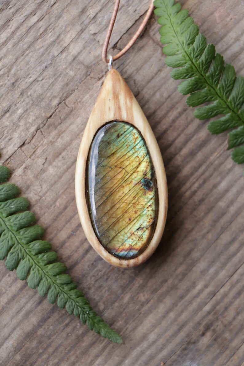 May include: A teardrop-shaped pendant featuring a polished, iridescent labradorite gemstone set within a smooth, light-colored wooden frame. The pendant hangs from a thin, tan leather cord, showcasing natural wood grain and golden-green stone hues.