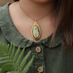 May include: A teardrop-shaped pendant necklace featuring a polished, iridescent green and gold labradorite stone set within a light-colored wooden frame, suspended from a thin brown cord worn around the neck against a dark green collared shirt.