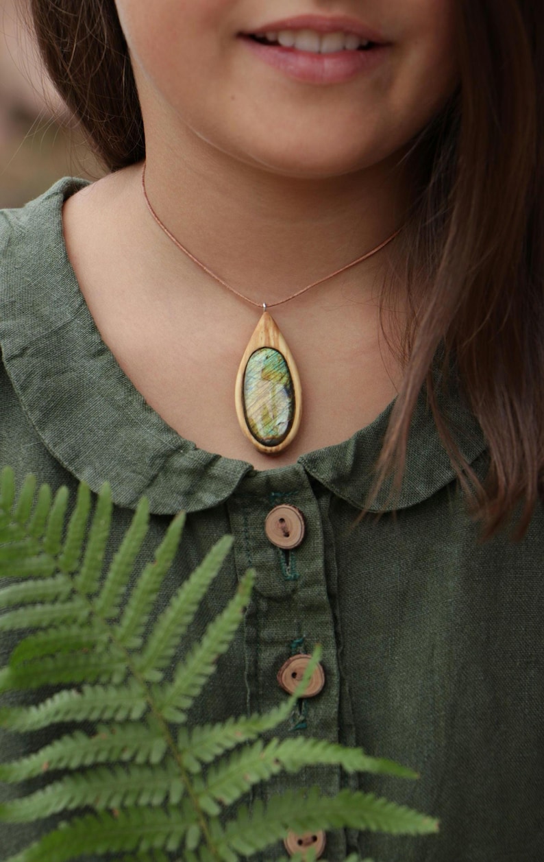 May include: A teardrop-shaped pendant necklace featuring a polished, iridescent green and gold labradorite stone set within a light-colored wooden frame, suspended from a thin brown cord worn around the neck against a dark green collared shirt.