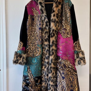 May include: A long coat featuring a patchwork design of magenta, teal, and black fabric panels with gold metallic brocade patterns and sequins. The coat has a leopard print faux fur trim along the front opening, the hood, and the sleeve cuffs.